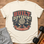Cream t-shirt with "BUILT DIFFERENT MOSTLY TIRED" graphic featuring a tired barbell character.