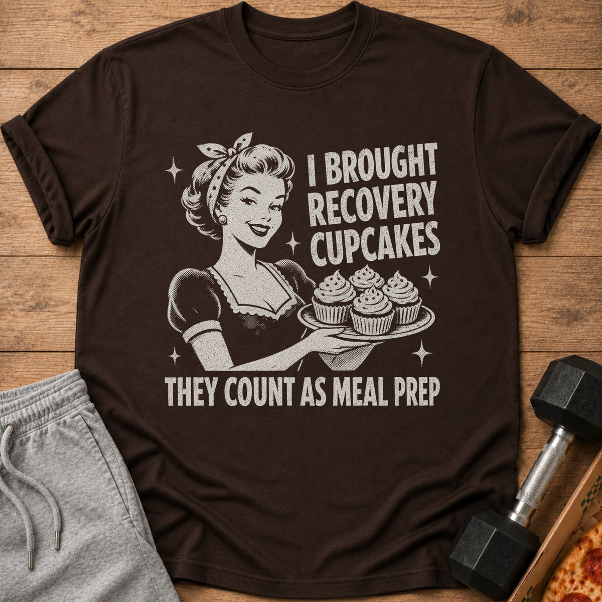 Brown t-shirt graphic of a woman holding cupcakes with text "I BROUGHT RECOVERY CUPCAKES THEY COUNT AS MEAL PREP".
