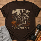Brown t-shirt with skeleton lifting weights and text "HAUNTED BY ONE MORE SET".