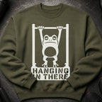 Olive green sweatshirt with a white graphic of a tired frog hanging from a bar. Text reads "HANGING IN THERE".