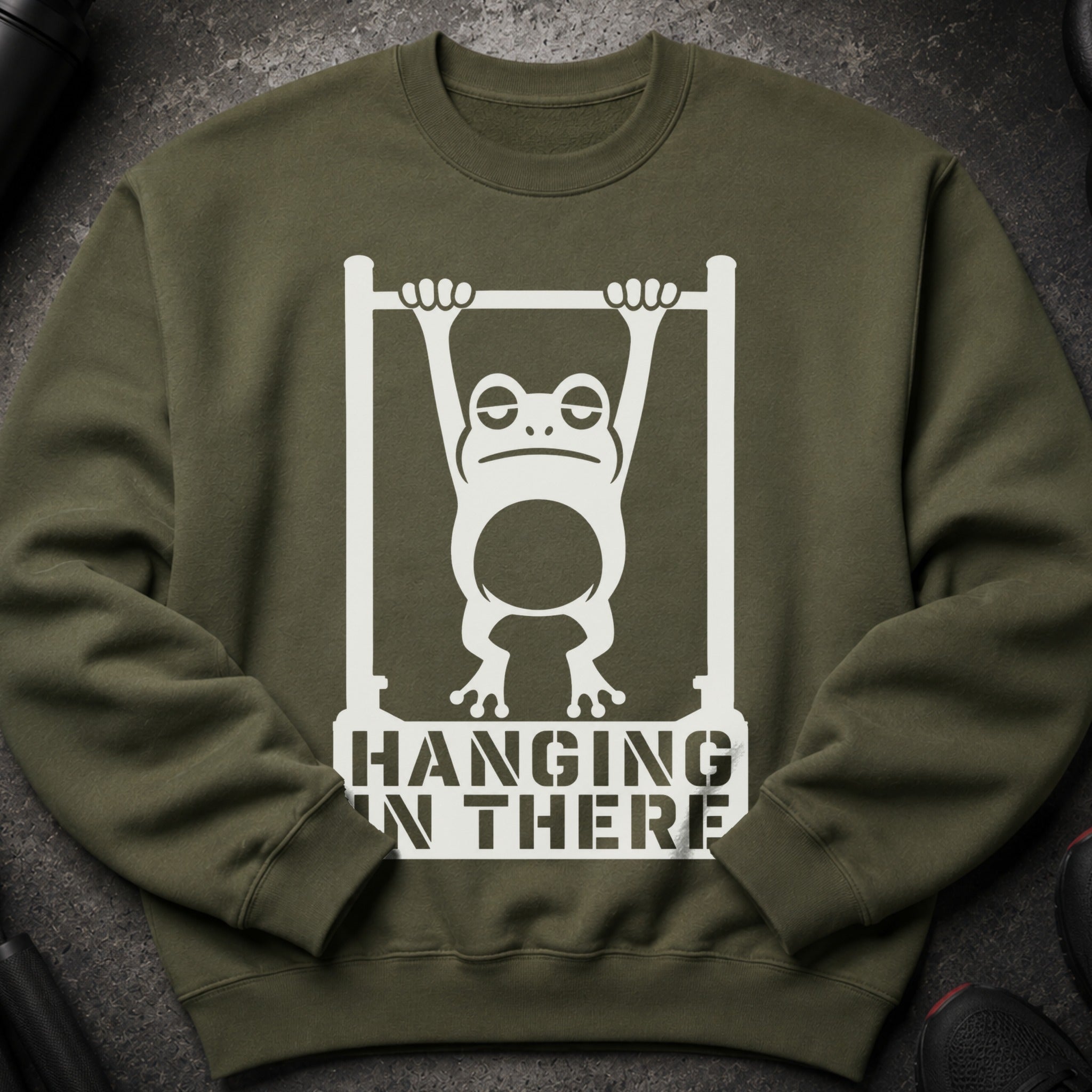 Olive green sweatshirt with a white graphic of a tired frog hanging from a bar. Text reads "HANGING IN THERE".