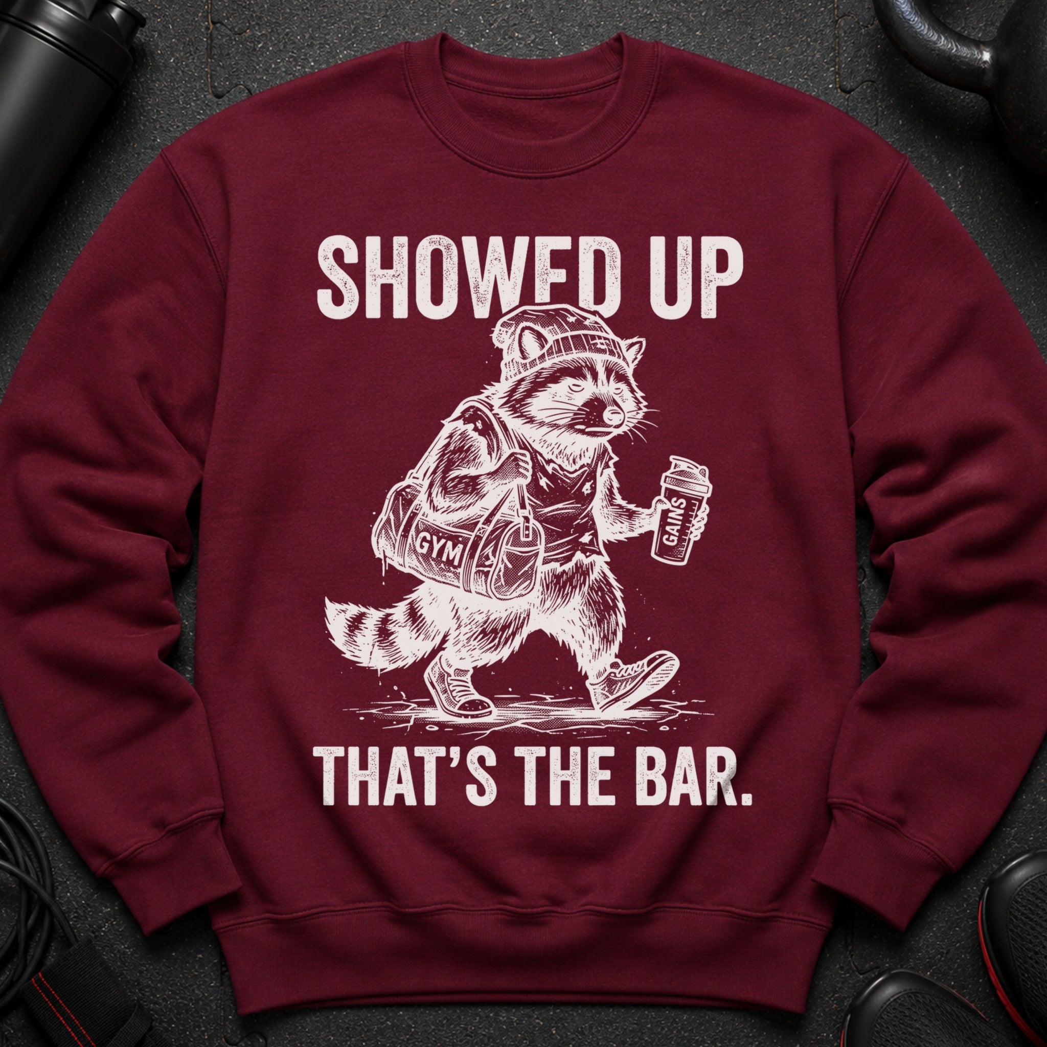 Maroon sweatshirt with graphic of a raccoon in gym clothes carrying a bag and shaker bottle.