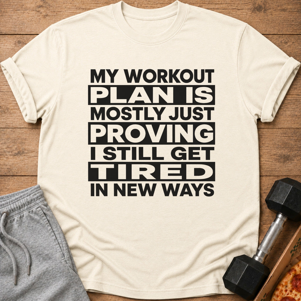 Light tan t-shirt with black text reading "MY WORKOUT PLAN IS MOSTLY JUST PROVING I STILL GET TIRED IN NEW WAYS".
