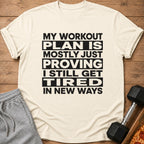 Light tan t-shirt with black text reading "MY WORKOUT PLAN IS MOSTLY JUST PROVING I STILL GET TIRED IN NEW WAYS".