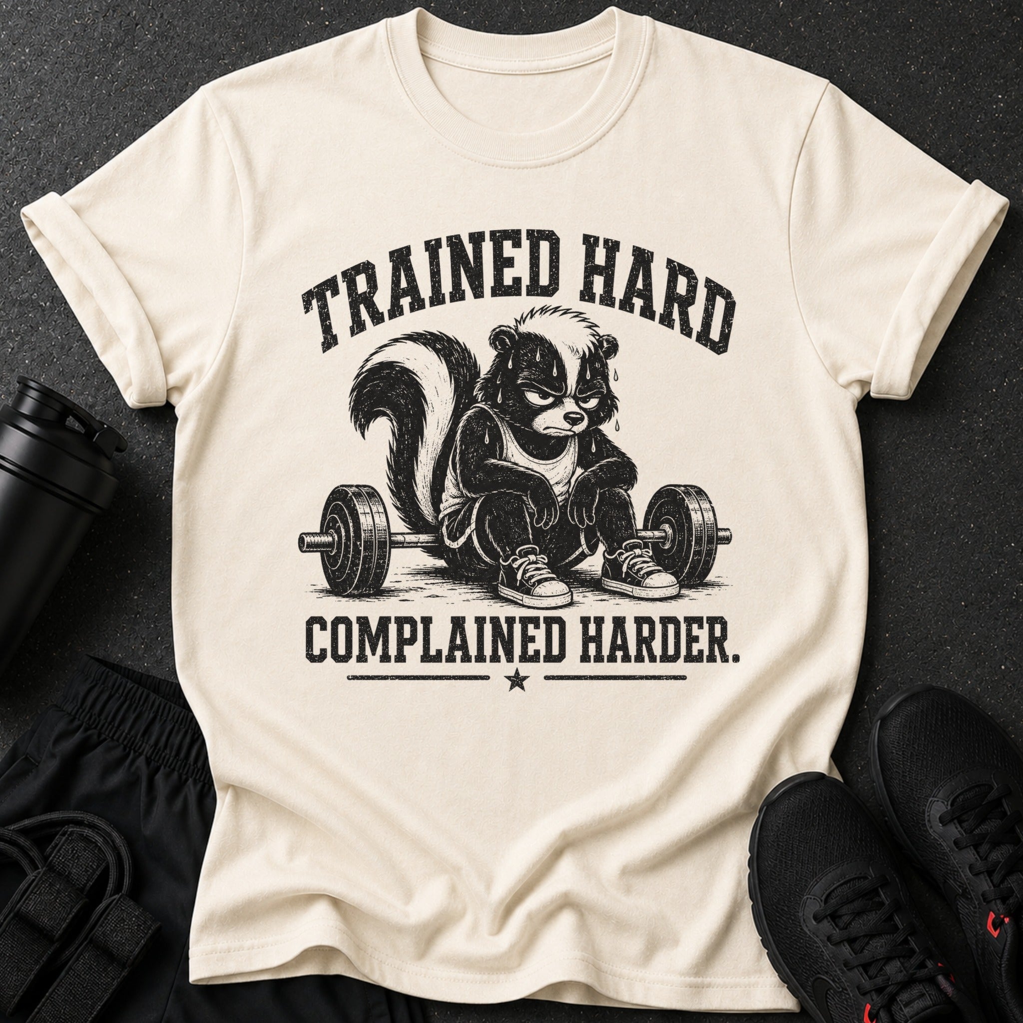 Trained hard complained harder graphic on a cream t-shirt with a skunk at a barbell.