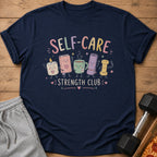 Navy t-shirt with "SELF-CARE STRENGTH CLUB" and cartoon candle, book, mug, yoga mat, bolt.