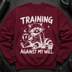 Maroon sweatshirt with a tired raccoon holding coffee and a dumbbell, text reads "TRAINING AGAINST MY WILL."