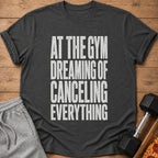 Dark gray t-shirt with white text: "AT THE GYM DREAMING OF CANCELING EVERYTHING".