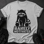 Grey t-shirt with a black graphic of a raccoon holding kettlebells and text reading "RARELY MOTIVATED".