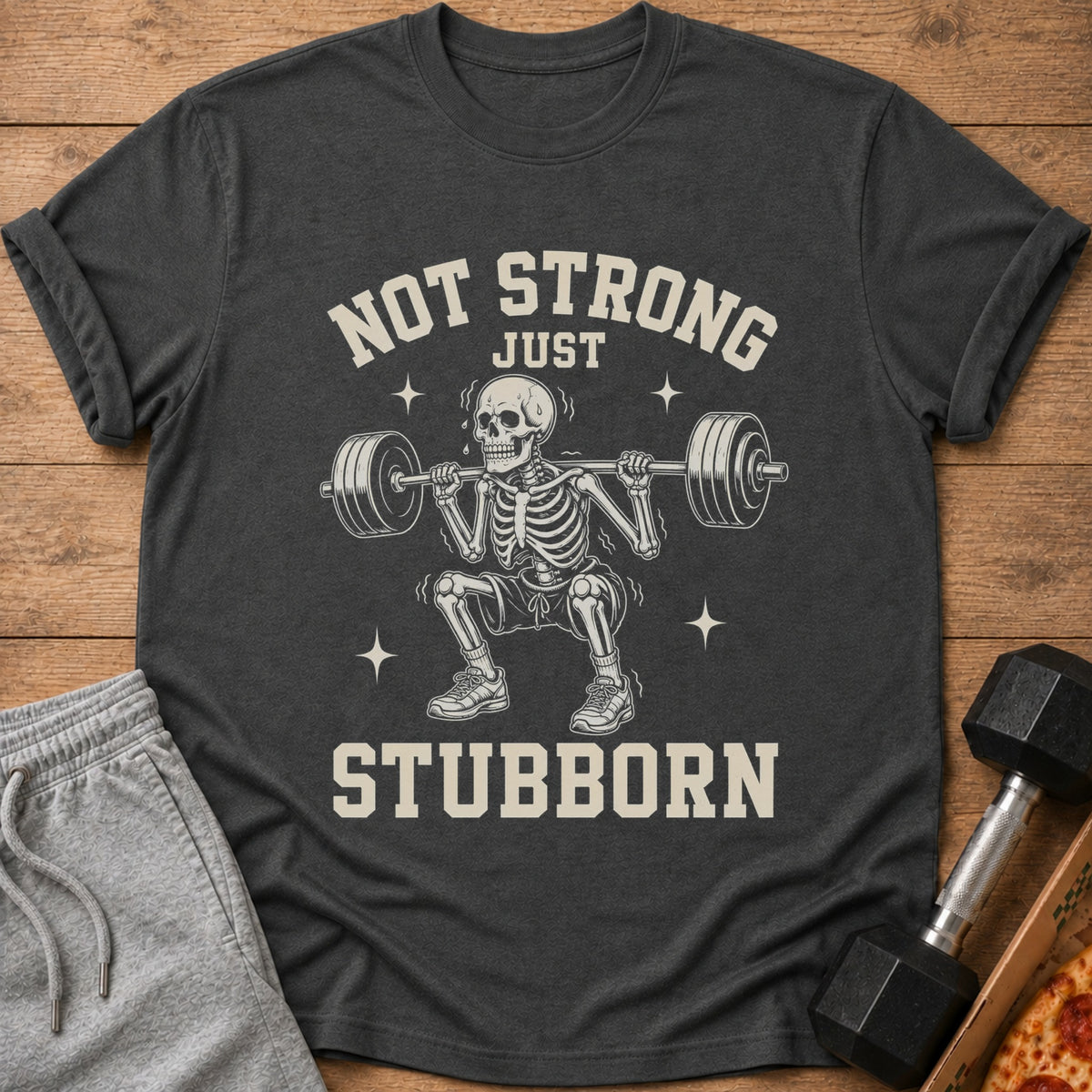 Dark grey t-shirt with skeleton lifting weights and text "NOT STRONG JUST STUBBORN".