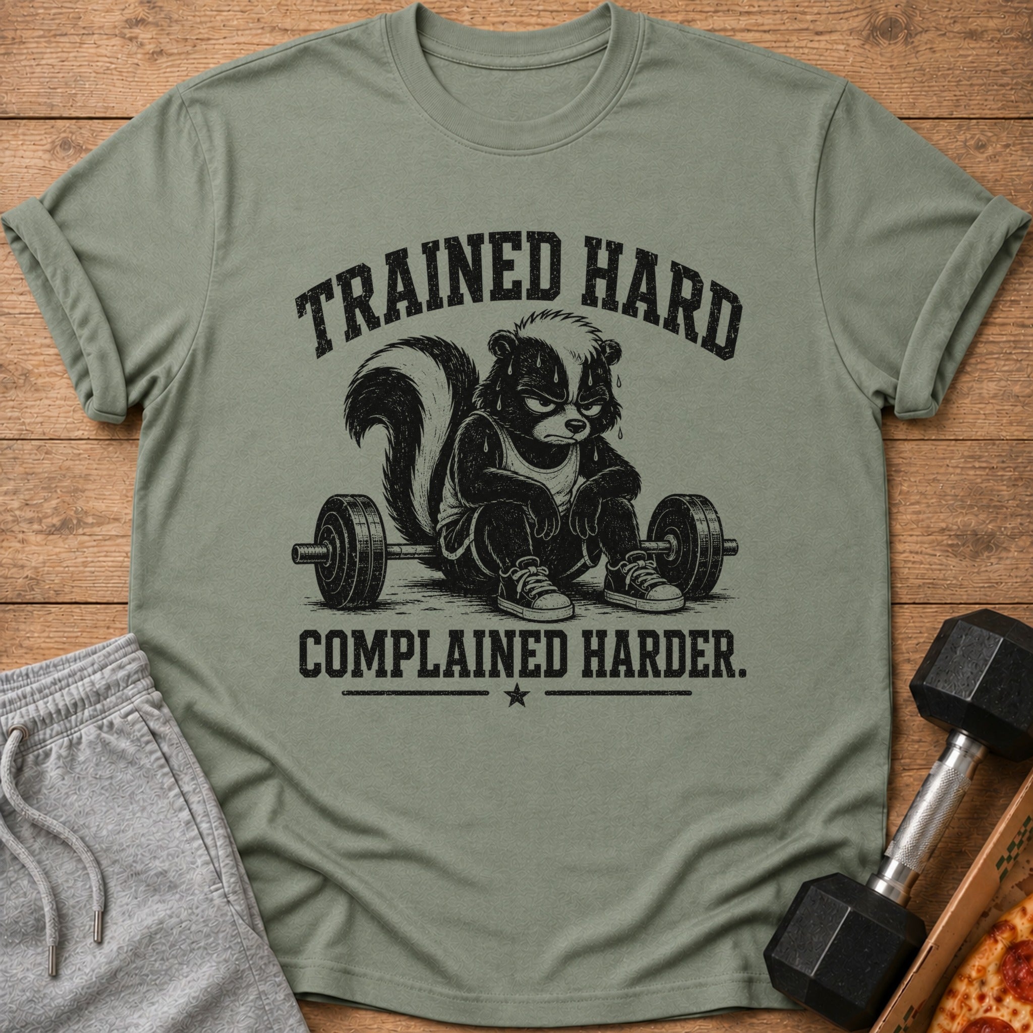 T-shirt graphic with a grumpy skunk sitting by a barbell, text reads "TRAINED HARD COMPLAINED HARDER."