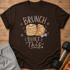 A brown crewneck t-shirt featuring a cartoon illustration of pancakes and a croissant flexing with dumbbells, with the text "BRUNCH BUILT THIS".