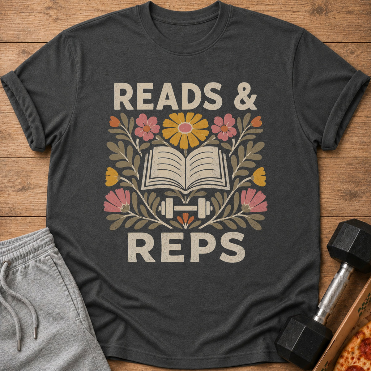 Dark gray t-shirt featuring a retro graphic with an open book, dumbbells, flowers, and the text READS & REPS.