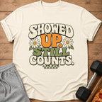 Cream t-shirt with "SHOWED UP. STILL COUNTS." in retro font and daisy graphics.
