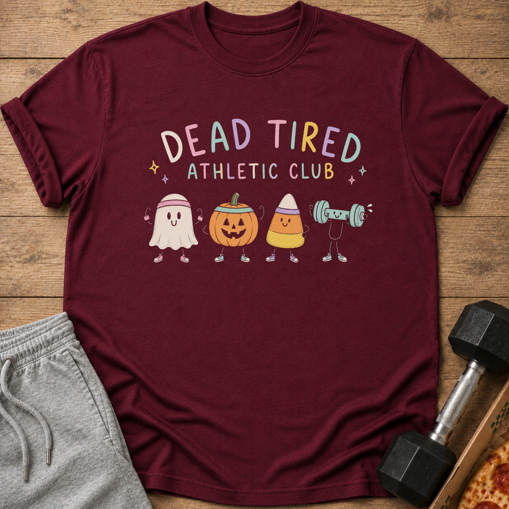 Maroon t-shirt with a "Dead Tired Athletic Club" graphic featuring cute Halloween characters lifting weights, laid on wood.