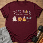 Maroon t-shirt with a "Dead Tired Athletic Club" graphic featuring cute Halloween characters lifting weights, laid on wood.