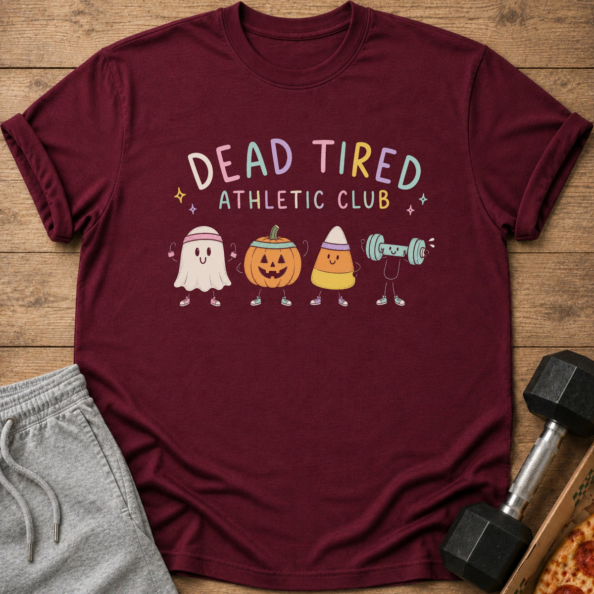 Maroon t-shirt with a "Dead Tired Athletic Club" graphic featuring cute Halloween characters lifting weights, laid on wood.
