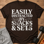 Brown t-shirt with "Easily Distracted By Snacks & Sets" text and illustrations.
