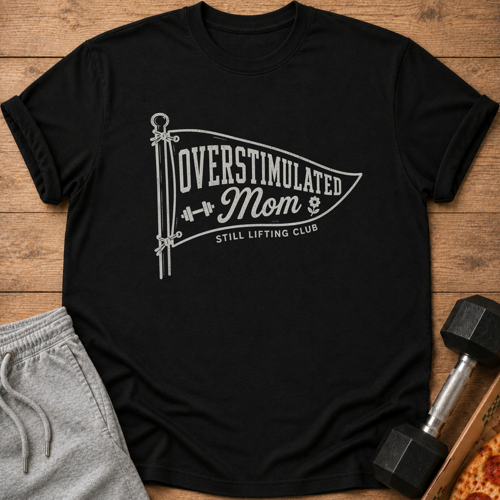 Black short-sleeved t-shirt with white graphic that reads Overstimulated Mom Still Lifting Club, displayed on wood.