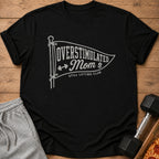 Black short-sleeved t-shirt with white graphic that reads Overstimulated Mom Still Lifting Club, displayed on wood.