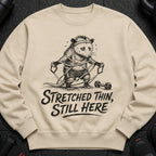 Sweatshirt with illustration of an exhausted opossum exercising, text reads "Stretched Thin, Still Here."
