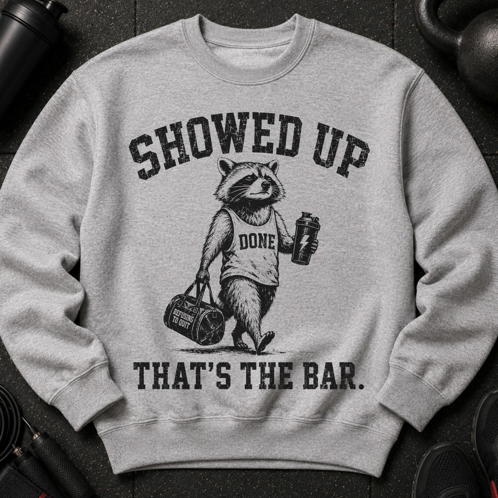 Grey graphic sweatshirt with raccoon holding gym bag and shaker, reading "Showed Up That's The Bar."