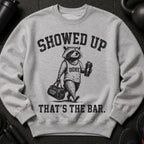 Grey graphic sweatshirt with raccoon holding gym bag and shaker, reading "Showed Up That's The Bar."
