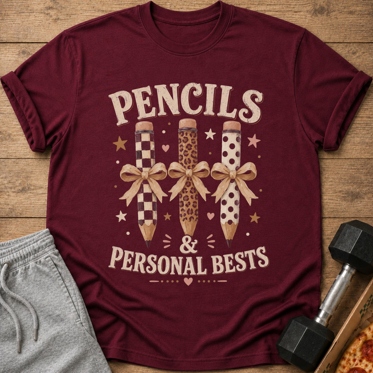 Maroon t-shirt with graphic of three pencils with patterns and bows, reading "Pencils & Personal Bests".