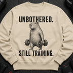 Cream sweatshirt with capybara holding weights and text "UNBOTHERED. STILL TRAINING."
