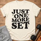 Cream t-shirt with "JUST ONE MORE SET" in black text, next to workout shorts and dumbbell.