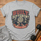 Gray t-shirt with "BUILT DIFFERENT MOSTLY TIRED" text and cartoon barbell character.