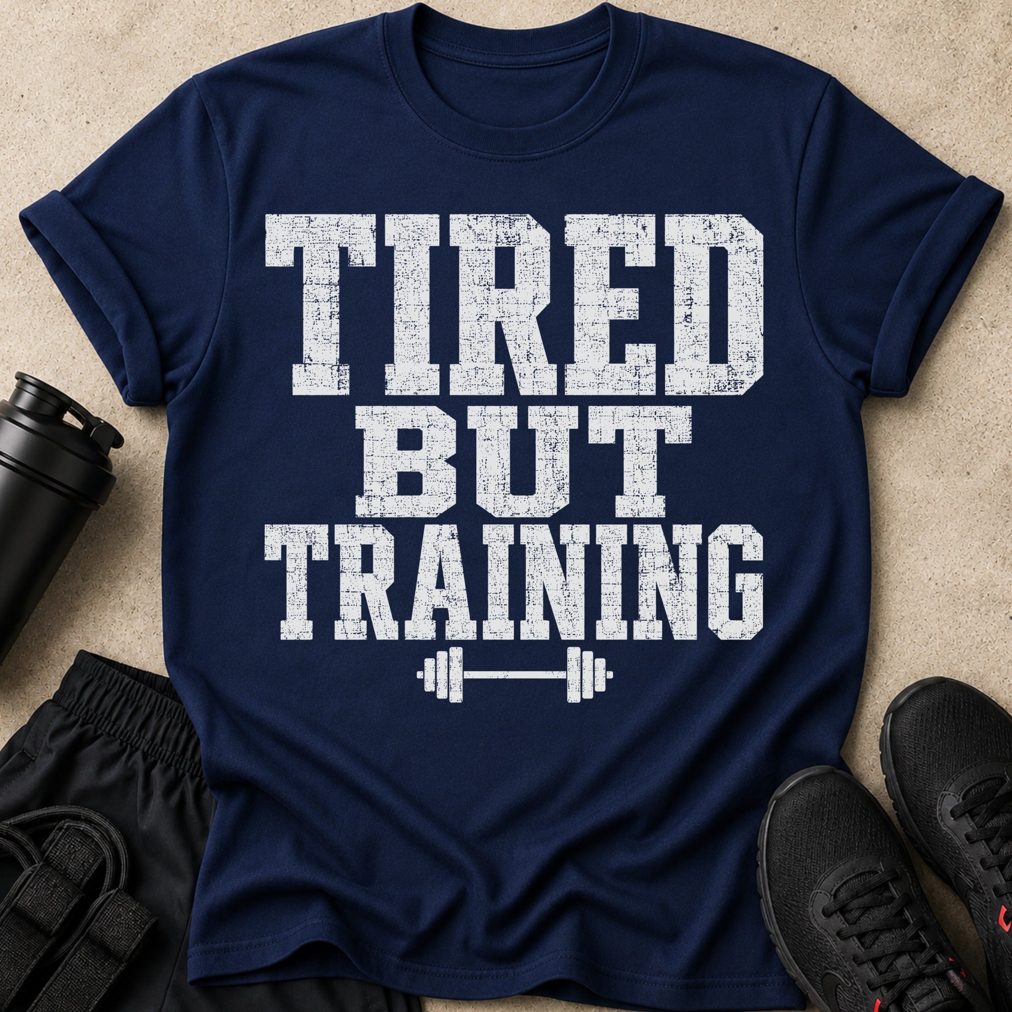 Navy t-shirt with white distressed text reading "TIRED BUT TRAINING" and a barbell graphic.