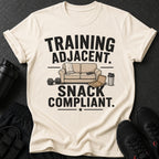 Cream t-shirt with black text "TRAINING ADJACENT. SNACK COMPLIANT." and graphics.