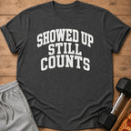 Dark t-shirt with white text "SHOWED UP STILL COUNTS" and gym accessories.