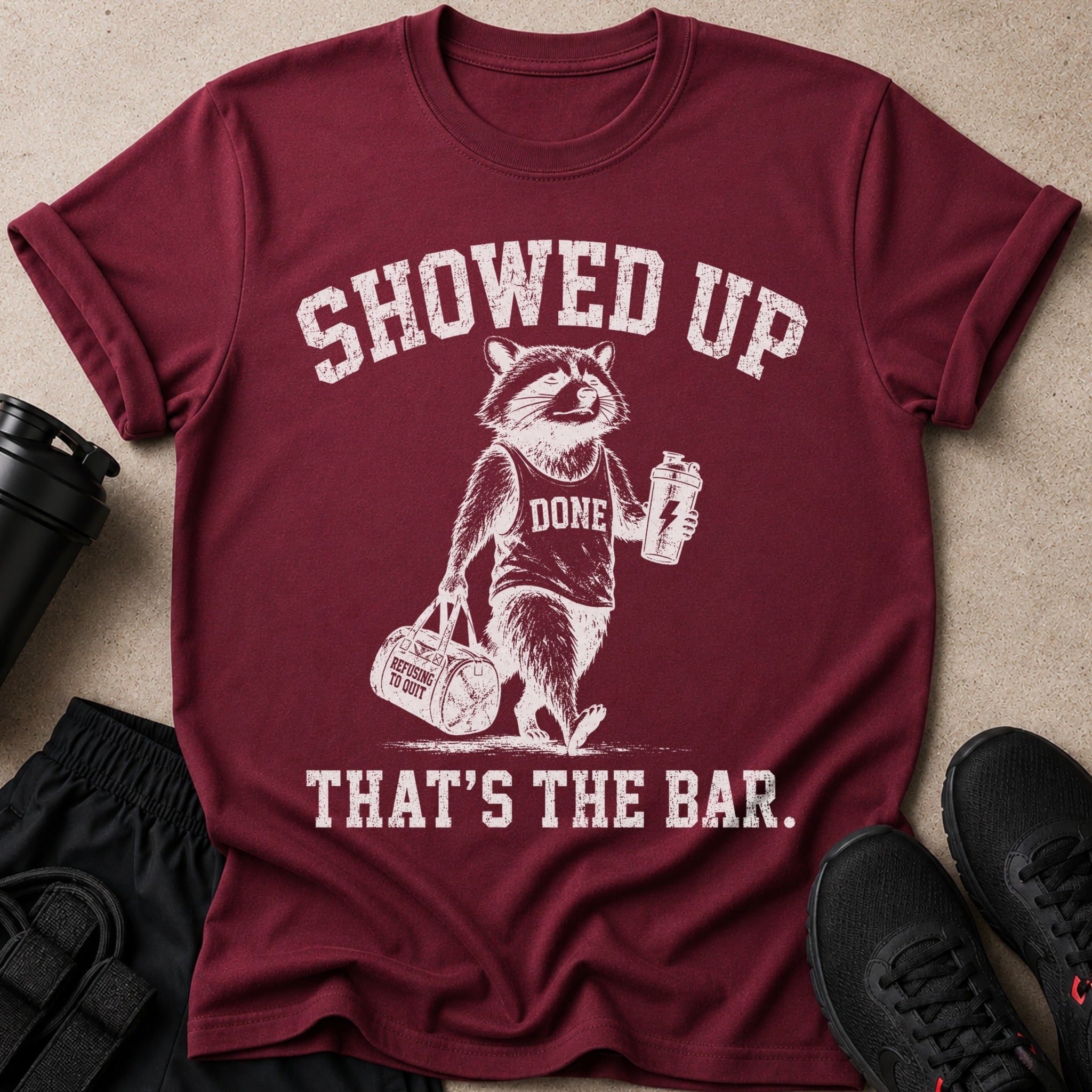 Maroon t-shirt with raccoon graphic saying "SHOWED UP DONE THAT'S THE BAR.".