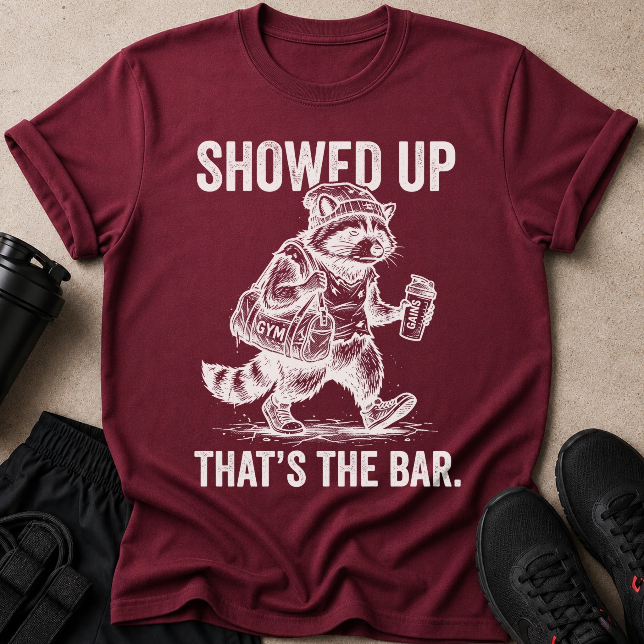 Maroon t-shirt with raccoon wearing hat, carrying gym bag, and protein shake.