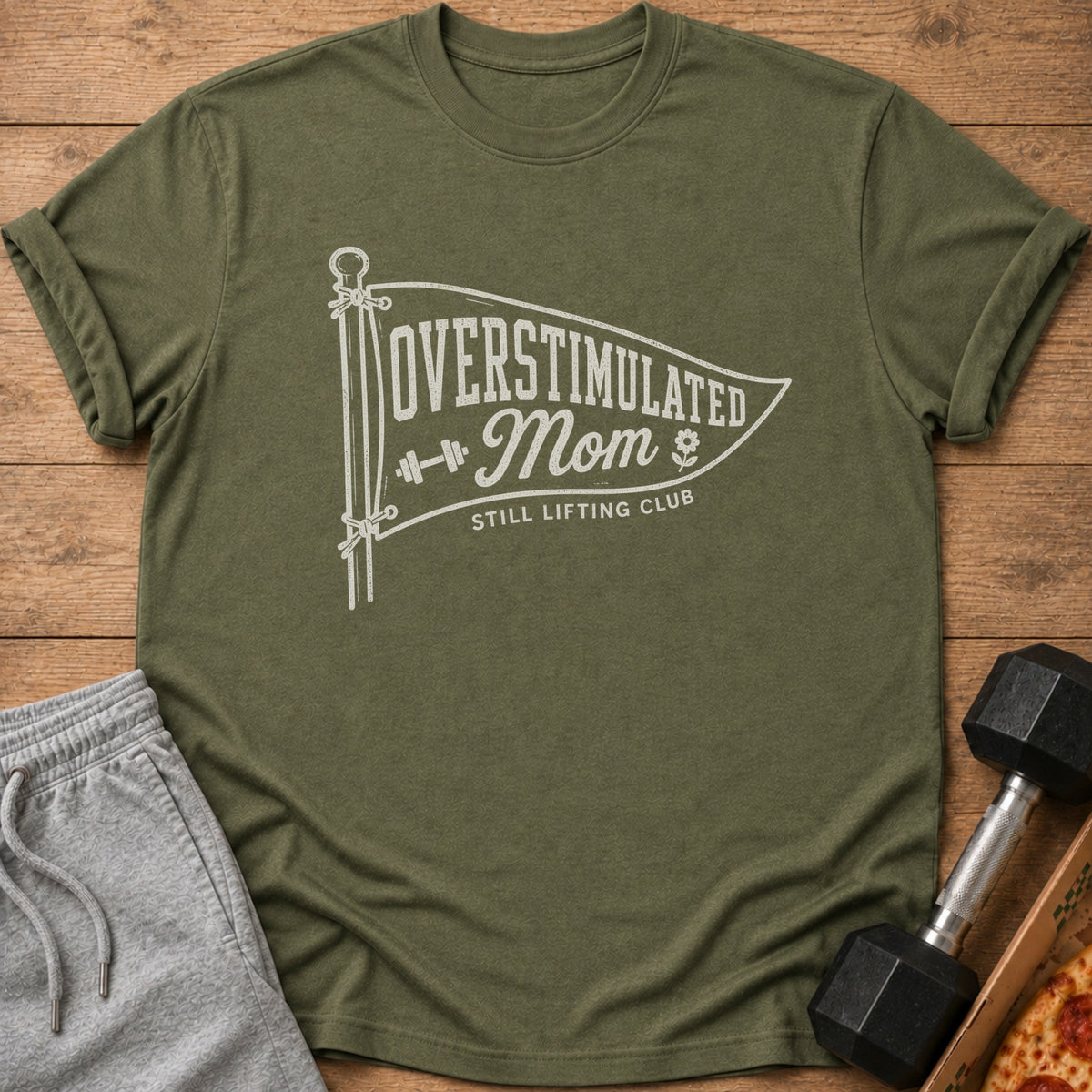 An olive green t-shirt with a white graphic featuring a pennant flag that reads OVERSTIMULATED Mom STILL LIFTING CLUB.