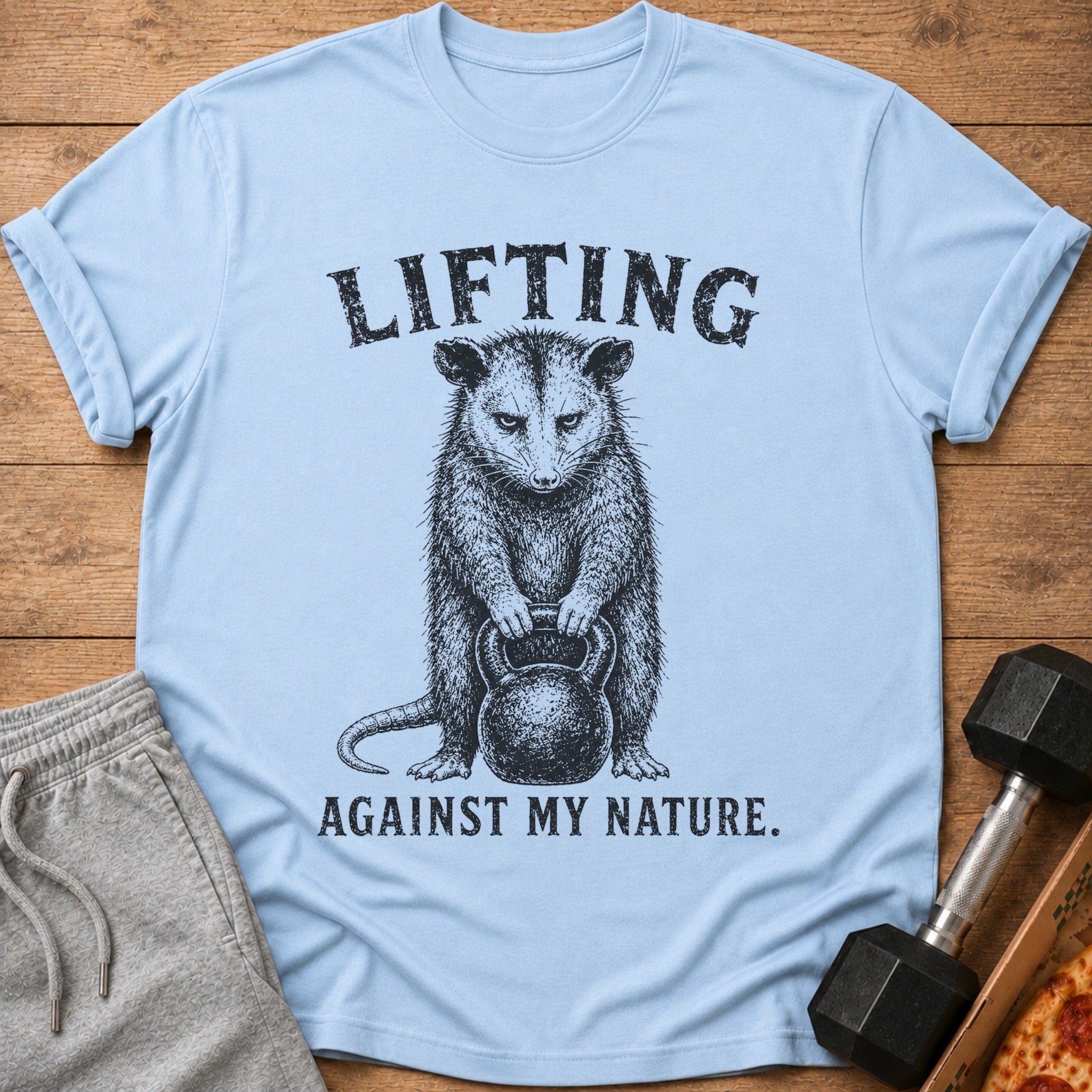 Light blue t-shirt with opossum lifting kettlebell, "LIFTING AGAINST MY NATURE." text.