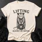 Cream t-shirt with opossum lifting kettlebell, text "LIFTING AGAINST MY NATURE."