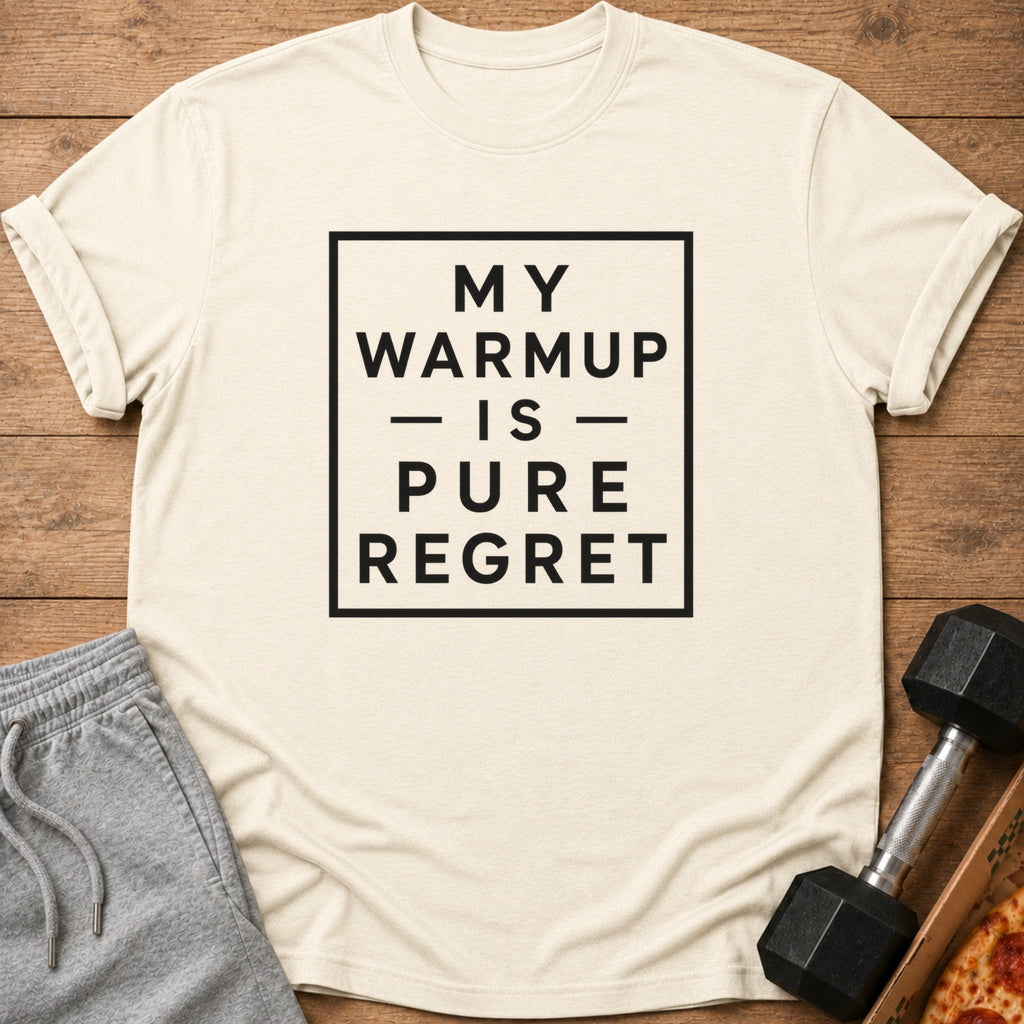 A beige t-shirt with black text reading "MY WARMUP IS PURE REGRET" lies on a wooden surface beside grey shorts and a dumbbell.