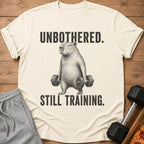 Cream t-shirt with capybara lifting dumbbells and text "UNBOTHERED. STILL TRAINING."