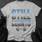 Gray t-shirt with faded blue and gray text "STILL COUNTS" above a barbell graphic.