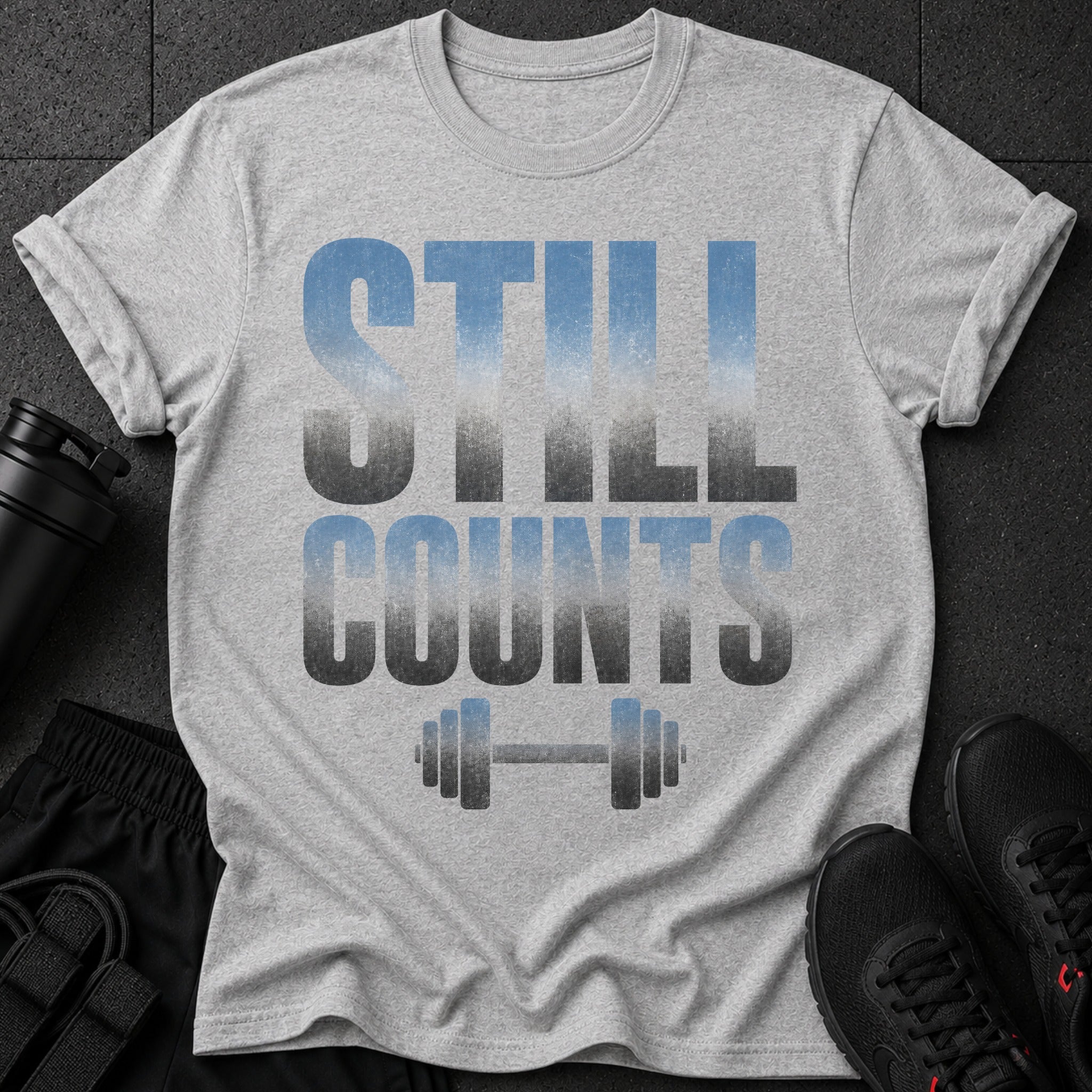 Gray t-shirt with faded blue and gray text "STILL COUNTS" above a barbell graphic.