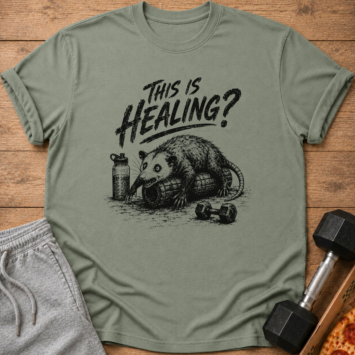 Green t-shirt with opossum on foam roller and text "This Is Healing?".