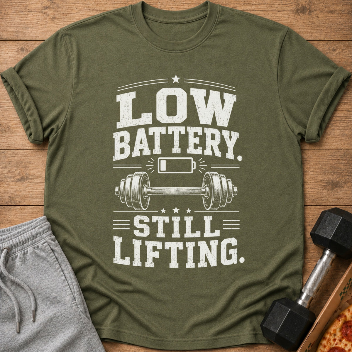 Olive green t-shirt with white text "LOW BATTERY. STILL LIFTING." and barbell graphic.