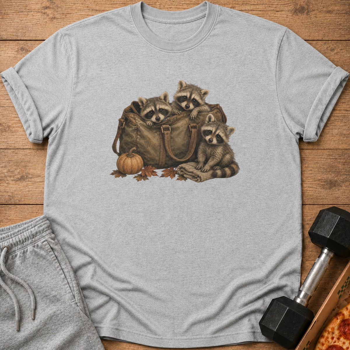 Three raccoons peek out of a duffel bag with a pumpkin and leaves.
