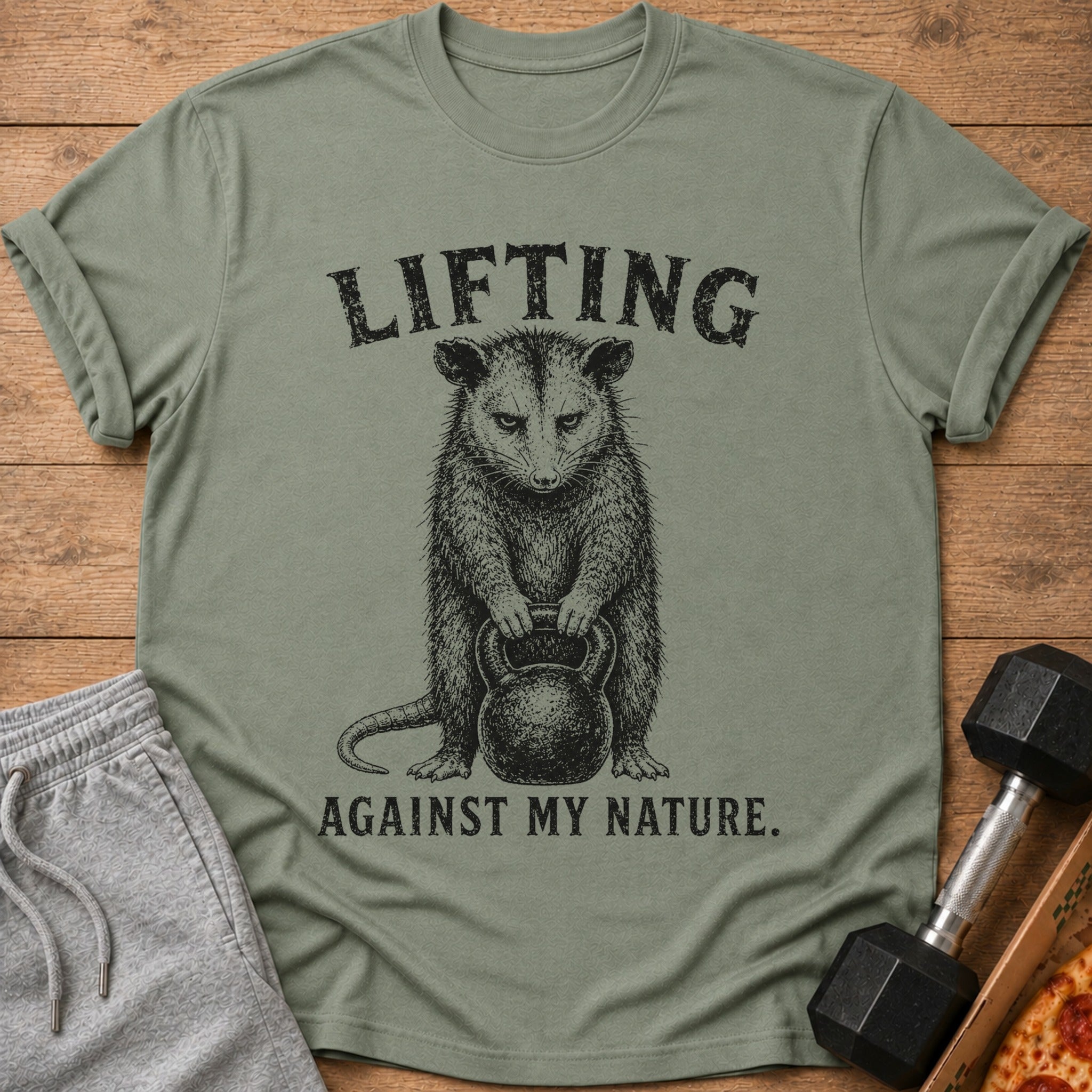 Green t-shirt with black text "LIFTING AGAINST MY NATURE" and a drawing of an opossum lifting a kettlebell.