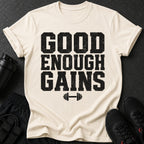 Cream t-shirt with black distressed text "GOOD ENOUGH GAINS" and dumbbell graphic.
