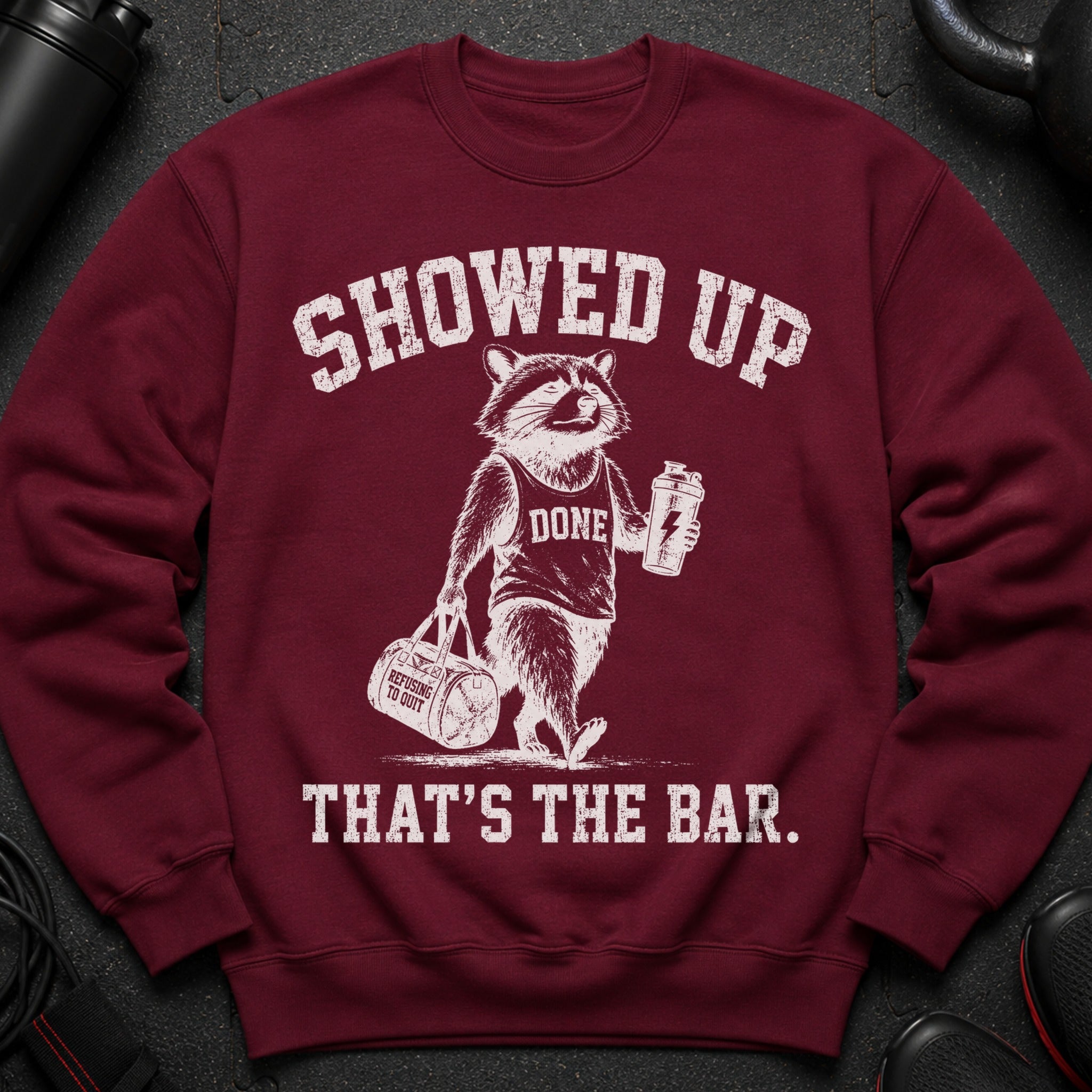 Maroon sweatshirt with a raccoon wearing a "DONE" tank top holding a shaker and gym bag.