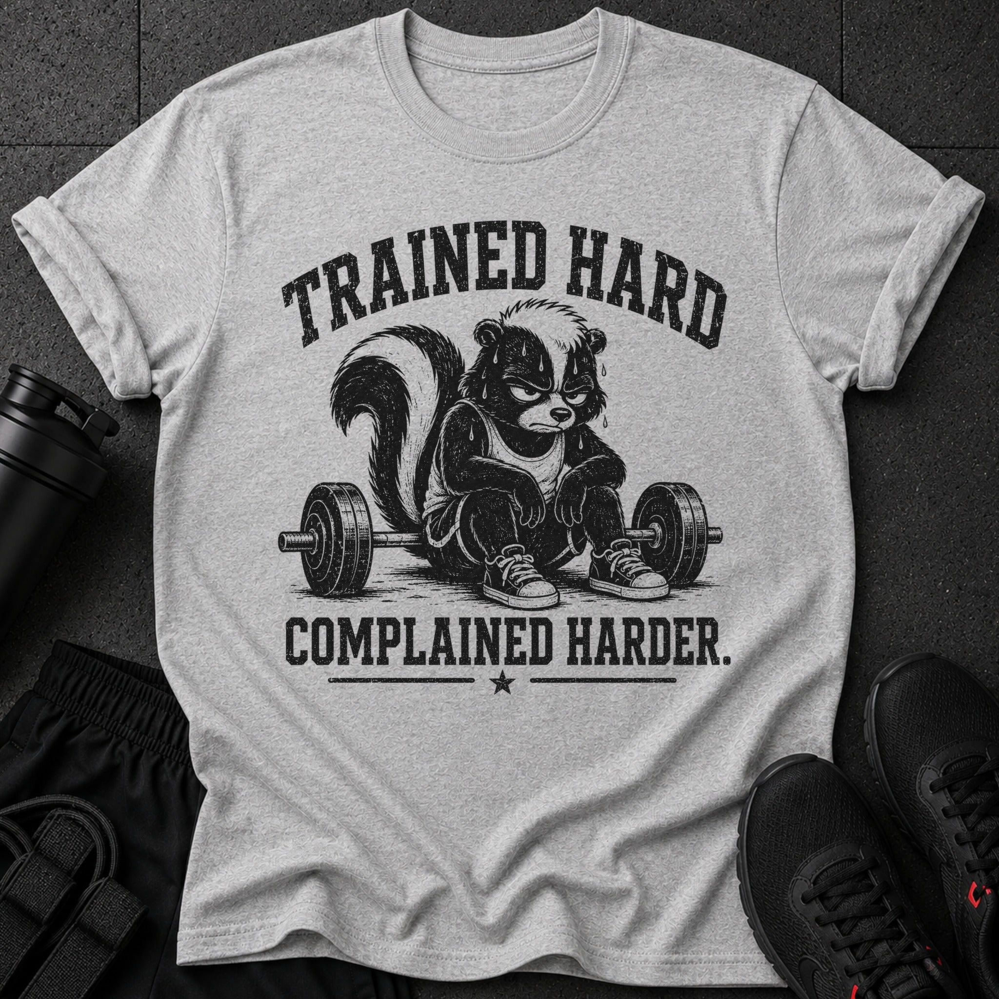 Grey t-shirt with a cartoon skunk sitting on a barbell, text reads "Trained Hard Complained Harder."